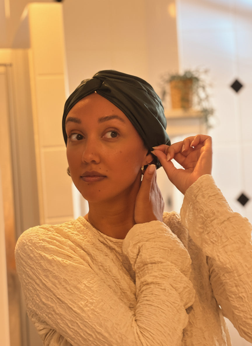 Vrouw zet een donkergroene zijden bonnet op voor de spiegel als onderdeel van haar avondritueel – natuurlijk haarverzorgmoment met Empowherment